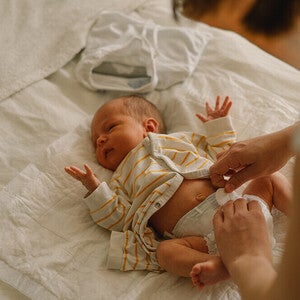 Photograph of newborn on white sheet in striped outfit, adult hands adjusting diaper
