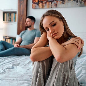Photograph of a pensive woman sitting on bed, distant man turned away