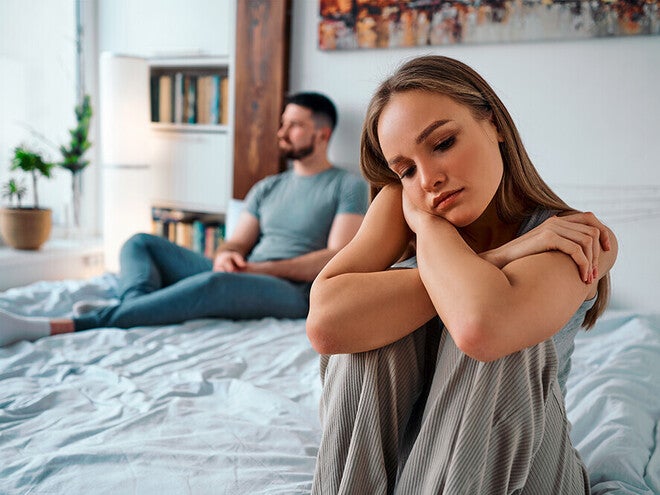 Photograph of a pensive woman sitting on bed, distant man turned away