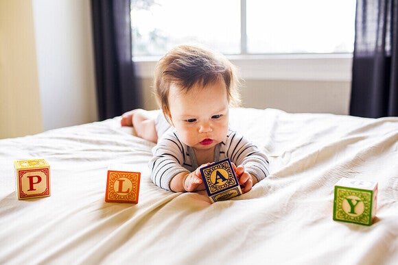 Photograph of a focused infant lying on a bed reaching for colorful wooden blocks
