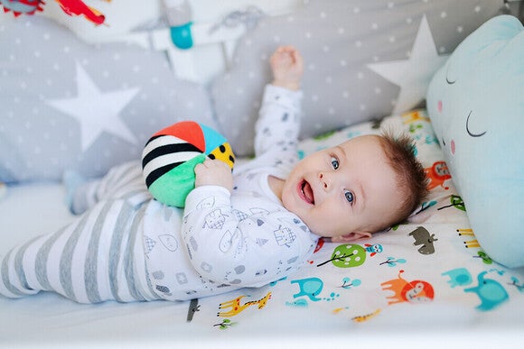 Smiling baby in crib with striped ball, cloud pillow, and star-pattern bedding.