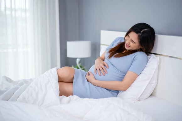 Photograph of a pregnant woman in blue dress reclining on white bed, cradling her belly.