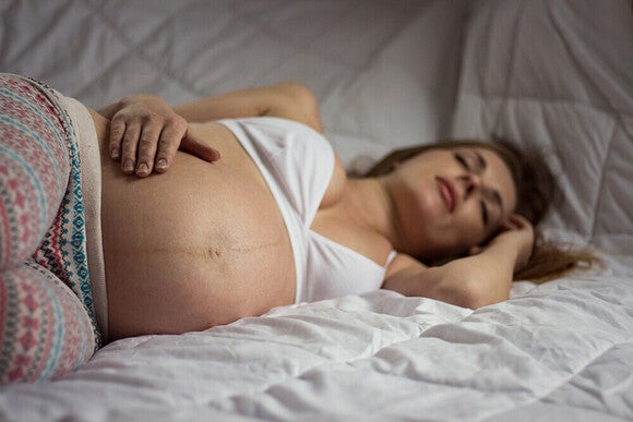 Photograph: pregnant woman reclining on bed, hand resting on bare belly, eyes closed