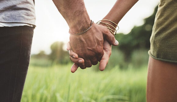 Photograph of two people holding hands in a grassy field at sunset, bracelets visible
