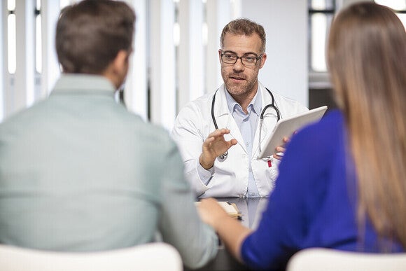 Photograph: male doctor consulting a seated couple, gesturing with clipboard.